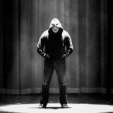 man standing on a stage with hands in his pocket wearing a hoodie